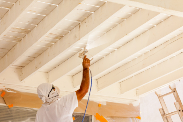 Professional Deck Painter underneath the deck with sprayer and mask on.